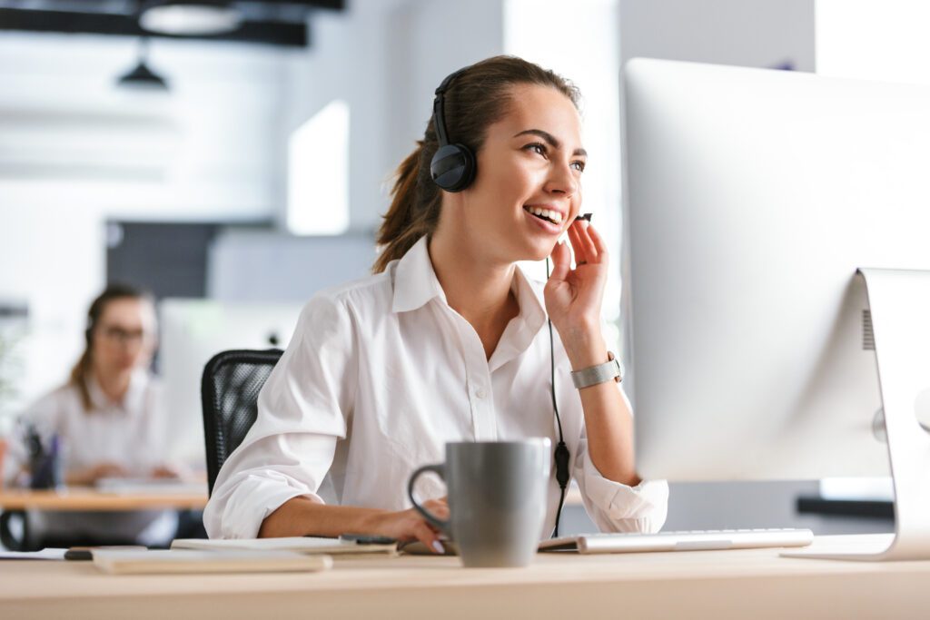 Image of a happy emotional business woman in office callcenter working with computer wearing headphones.