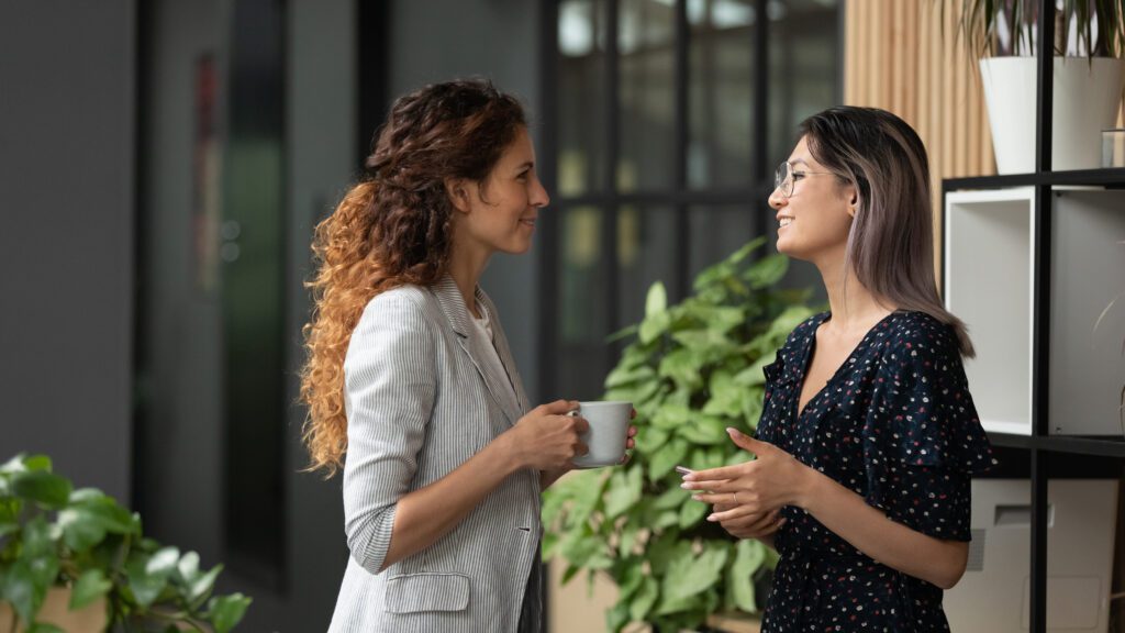 Smiling millennial multiracial female employees talk chat during coffee break in office, young multiethnic women colleagues speak laughing enjoy leisure time at workplace, cooperating concept