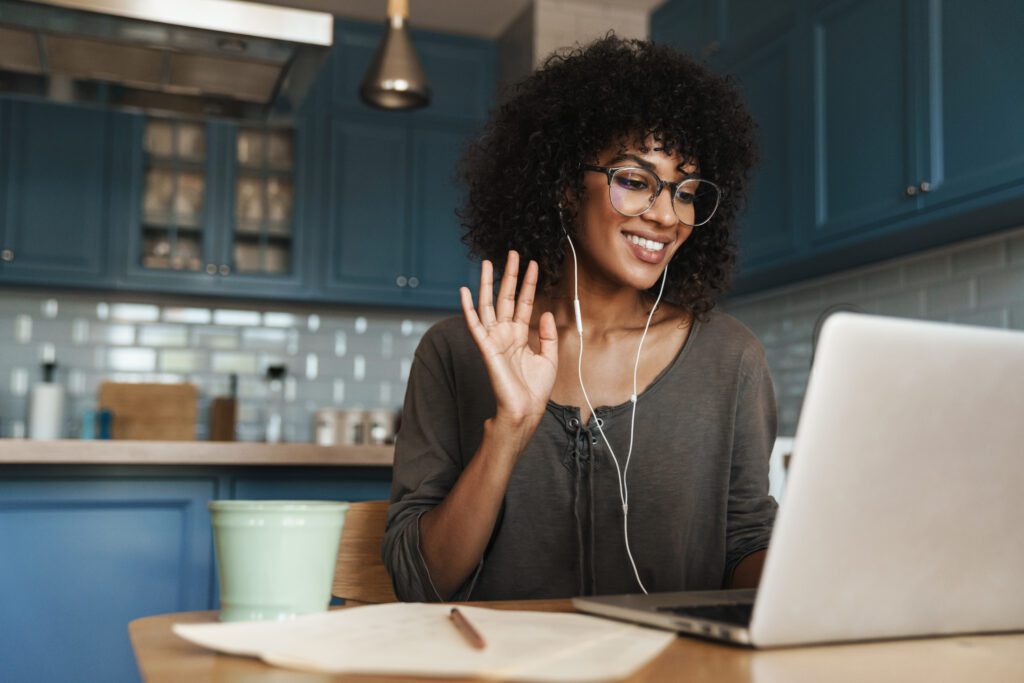 Attractive smiling young african woman on a video call with colleagues, sitting on a kitchen with laptop computer, waving