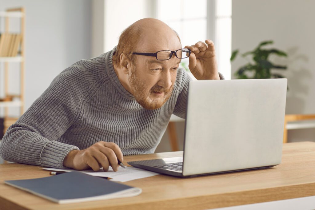 Eyesight problems. Confused mature man who has poor eyesight and works on laptop at home wearing glasses. Elderly man lifts his glasses from his eyes and squints while looking at computer screen.