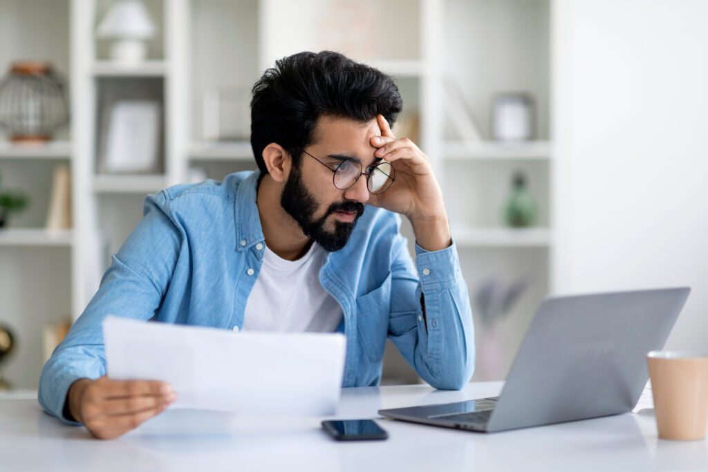 Stressed Young Indian Man Looking At Laptop Screen And Holding Documents, Focused Millennial Eastern Male Cheching Business Reports While Working Remotely At Home Office, Closeup Shot With Free Space