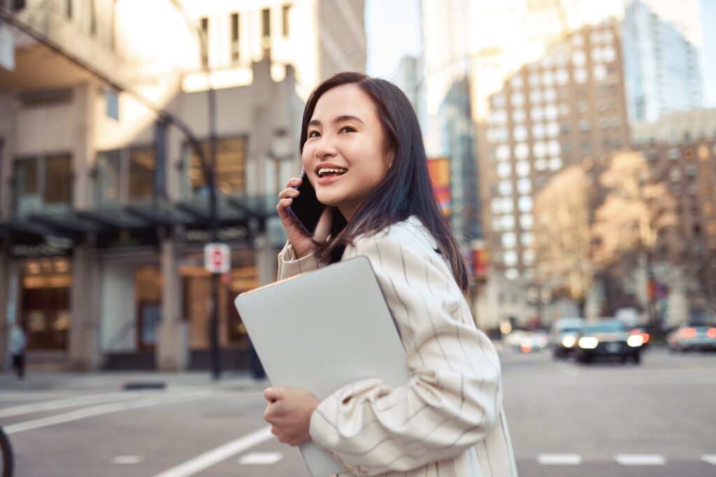 Young busy happy Asian business woman office professional holding cellphone in hands walking on big city urban street making corporate business call, talking on the cellular phone. Authentic shot