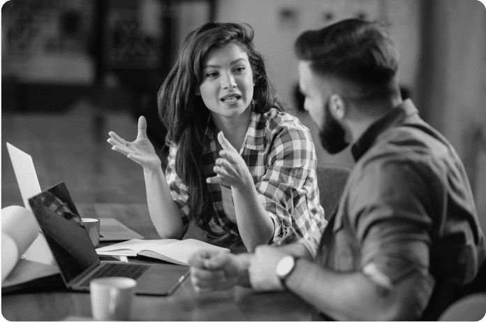 Woman in a plaid shirt explains something to a man in front of a computer.