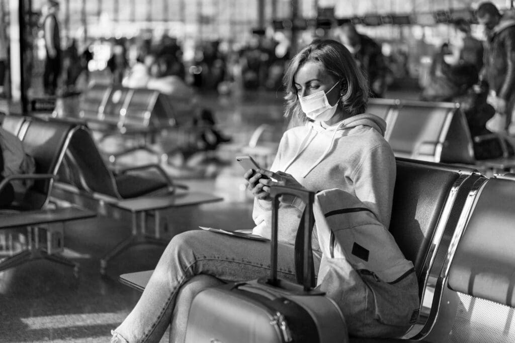 A European woman in a protective medical mask on her face sits at the airport waiting for a flight and uses the phone.
